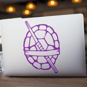 Könnte beinhalten: Ein violetter Aufkleber mit einem Schildkrötenpanzer, zwei gekreuzten Schwertern und einem Stirnband, der auf einem weißen Laptop angebracht ist. Der Laptop steht auf einem Holztisch.