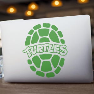 May include: A white laptop with a green turtle shell design and the word "TURTLES" in a matching green font. The design is centered on the laptop's lid. The laptop is on a wooden surface.