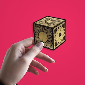 Op de afbeelding: Een zwarte en gouden puzzelkubus met ingewikkelde geometrische patronen. De kubus wordt tegen een felroze achtergrond gehouden. De doos heeft een groot cirkelvormig ontwerp aan één kant en andere gedetailleerde patronen.