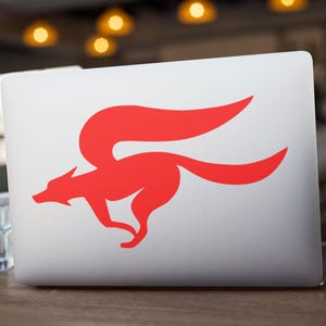 Könnte beinhalten: Ein silberner Laptop mit einem roten Fuchslogo auf dem Deckel, auf einem Holztisch. Das Fuchsmotiv ist stilisiert und scheint sich zu bewegen. Links vom Laptop steht ein Glas Wasser.