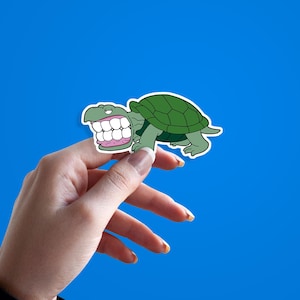 May include: A cartoon sticker of a green turtle with a large, toothy grin. The turtle has a green shell and is set against a bright blue background. The sticker is held up by a hand.
