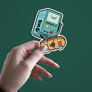 May include: A sticker featuring a cartoon character, a teal-colored video game console with a smiling face, riding a skateboard. The skateboard is orange with a skull design. The sticker has a white border and the letters "BMO" are visible.