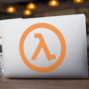 May include: A white laptop computer with an orange lambda symbol sticker on the lid. The laptop is open and resting on a wooden table. A glass of water sits to the left of the laptop.
