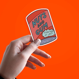 May include: A sticker featuring a cartoon-style can of "Nuts and Gum" in red and blue, with the text "TOGETHER AT LAST!" on a light blue banner. The sticker is held against an orange background.