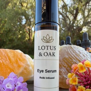 Może przedstawiać: Brązowa szklana butelka z czarną nakrętką, z napisem "LOTUS & OAK Eye Serum Reiki Infused". Butelka zawiera 10 ml / 0,34 fl oz. Tło obejmuje pomarańczowe i ż&oacute;łte kryształy, fioletowe i ż&oacute;łte kwiaty oraz rozmyte zielone drzewo.