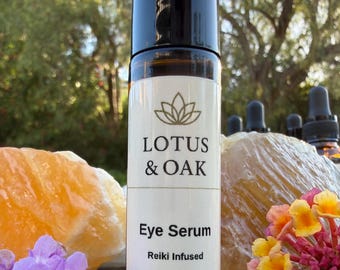 May include: A brown glass bottle with a black cap, labelled "LOTUS & OAK Eye Serum Reiki Infused." The bottle contains 10 ml / 0.34 fl oz. The background includes orange and yellow crystals, purple and yellow flowers, and a blurred green tree.