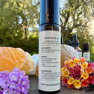 Może przedstawiać: Przezroczysta szklana butelka roll-on z czarną nakrętką, oznaczona jako "Botanical No. 2". Etykieta zawiera składniki i instrukcje. Butelka otoczona jest kwiatami, kryształami i innymi butelkami z olejkami eterycznymi.