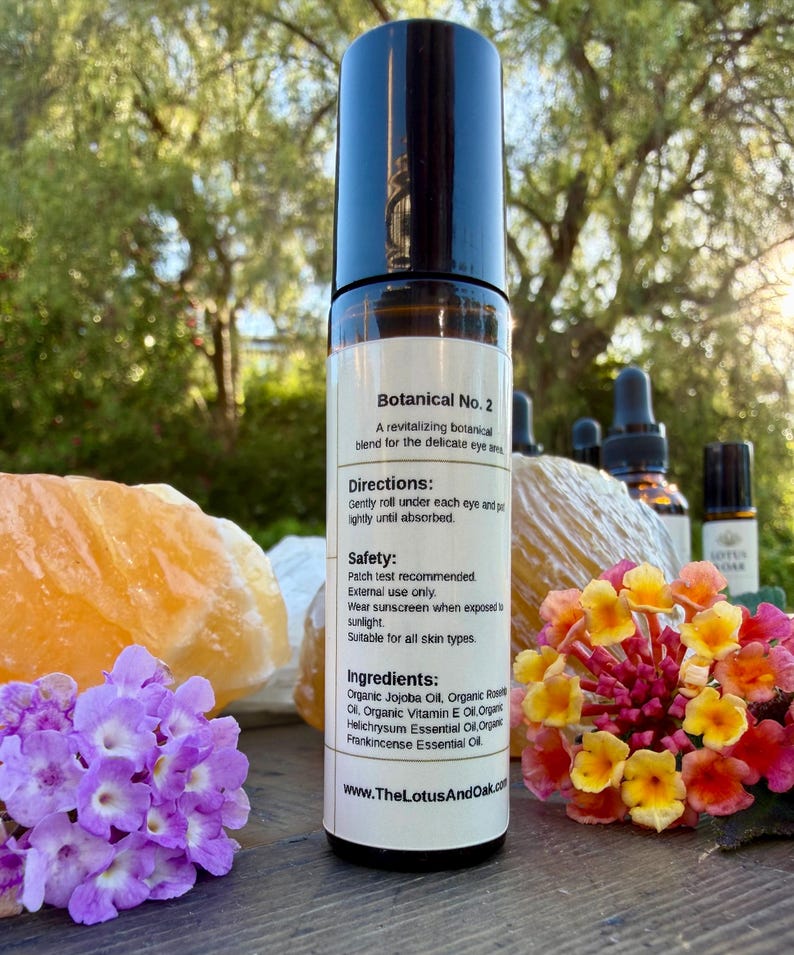 Może przedstawiać: Przezroczysta szklana butelka roll-on z czarną nakrętką, oznaczona jako "Botanical No. 2". Etykieta zawiera składniki i instrukcje. Butelka otoczona jest kwiatami, kryształami i innymi butelkami z olejkami eterycznymi.