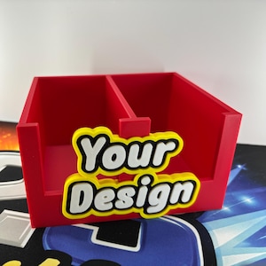 May include: A red desk tidy with two sections, featuring the words "Your Design" in white letters with yellow and black outlines. The organiser is made of a solid material and sits on a patterned surface.