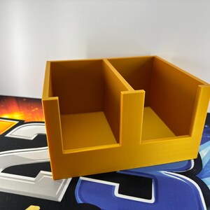 May include: A mustard-yellow, two-compartment organiser. The rectangular container has a divider creating two equal sections. The open-top design allows easy access to contents. The organiser is made of a solid material and sits on a patterned surface.