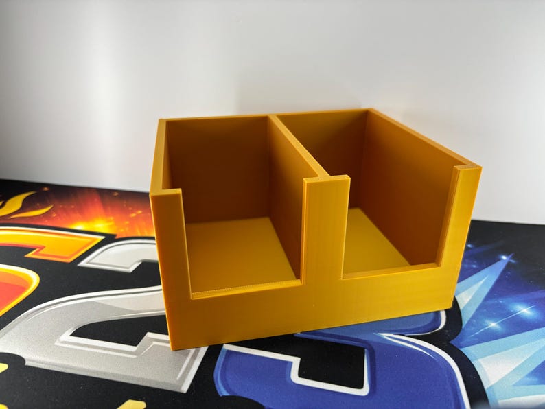 May include: A mustard-yellow, two-compartment organiser. The rectangular container has a divider creating two equal sections. The open-top design allows easy access to contents. The organiser is made of a solid material and sits on a patterned surface.