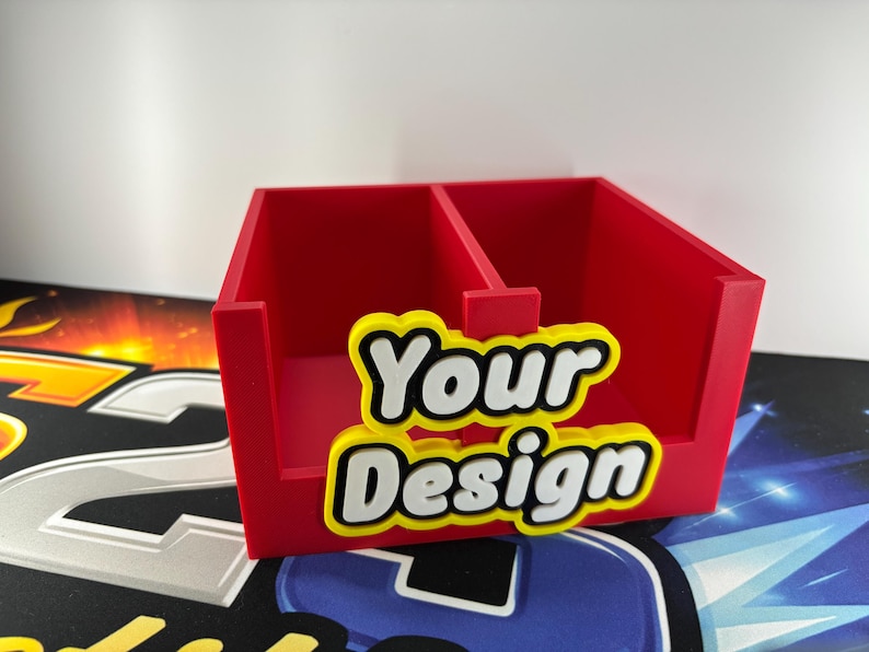 May include: A red desk tidy with two sections, featuring the words "Your Design" in white letters with yellow and black outlines. The organiser is made of a solid material and sits on a patterned surface.