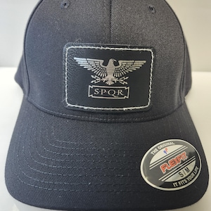 Peut inclure: Casquette de baseball Flexfit noire avec une visière incurvée. Le chapeau présente un écusson rectangulaire avec un aigle argenté et les lettres "SPQR". Un autocollant Flexfit est sur la visière.