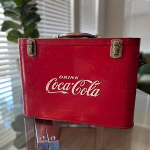 K&ouml;nnte beinhalten: Eine rote Vintage-Coca-Cola-K&uuml;hltasche mit braunem Griff und Metallverschl&uuml;ssen. Das wei&szlig;e Coca-Cola-Logo ist prominent auf der Vorderseite zu sehen. Die K&uuml;hltasche ist rechteckig und scheint aus Metall zu sein.