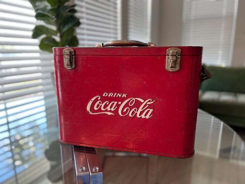 K&ouml;nnte beinhalten: Eine rote Vintage-Coca-Cola-K&uuml;hltasche mit braunem Griff und Metallverschl&uuml;ssen. Das wei&szlig;e Coca-Cola-Logo ist prominent auf der Vorderseite zu sehen. Die K&uuml;hltasche ist rechteckig und scheint aus Metall zu sein.