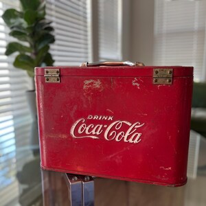 K&ouml;nnte beinhalten: Eine rote Vintage-Coca-Cola-K&uuml;hltasche mit dem wei&szlig;en Coca-Cola-Logo. Die K&uuml;hltasche hat einen Metallgriff und Scharniere und weist Gebrauchsspuren auf. Die K&uuml;hltasche ist rechteckig und scheint aus Metall zu sein.