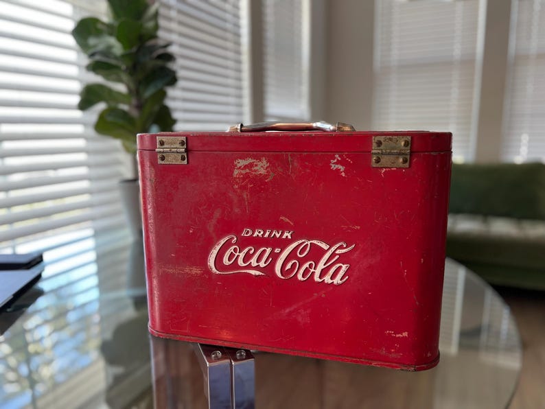 K&ouml;nnte beinhalten: Eine rote Vintage-Coca-Cola-K&uuml;hltasche mit dem wei&szlig;en Coca-Cola-Logo. Die K&uuml;hltasche hat einen Metallgriff und Scharniere und weist Gebrauchsspuren auf. Die K&uuml;hltasche ist rechteckig und scheint aus Metall zu sein.