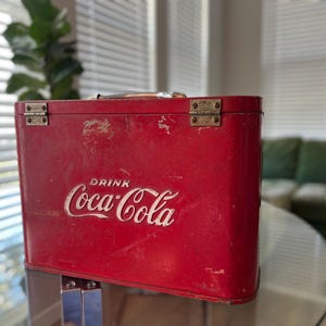 K&ouml;nnte beinhalten: Vintage rote Coca-Cola-K&uuml;hltasche mit dem wei&szlig;en Schriftzug "DRINK Coca-Cola". Die K&uuml;hltasche hat eine rechteckige Form, Metallbeschl&auml;ge und einen Griff. Die K&uuml;hltasche ist wahrscheinlich aus Metall und weist Gebrauchsspuren auf.