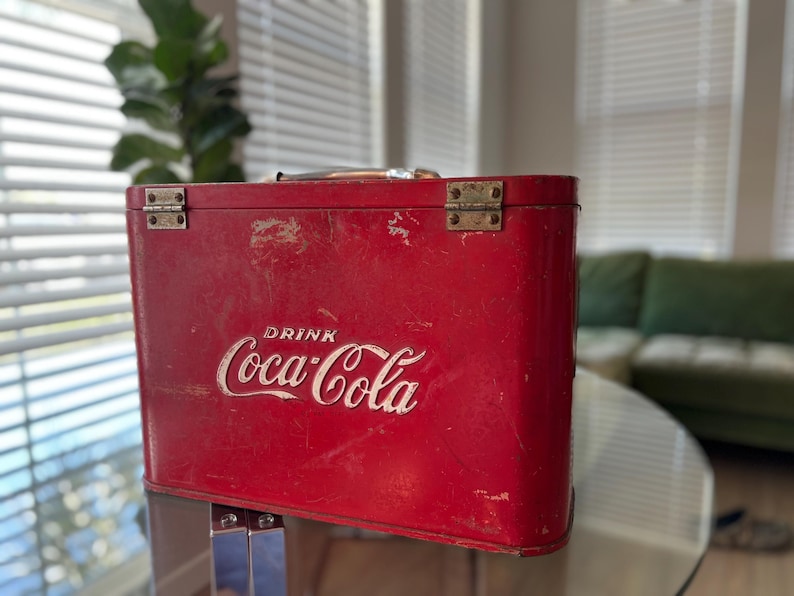 K&ouml;nnte beinhalten: Vintage rote Coca-Cola-K&uuml;hltasche mit dem wei&szlig;en Schriftzug "DRINK Coca-Cola". Die K&uuml;hltasche hat eine rechteckige Form, Metallbeschl&auml;ge und einen Griff. Die K&uuml;hltasche ist wahrscheinlich aus Metall und weist Gebrauchsspuren auf.