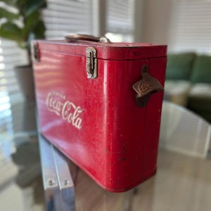 K&ouml;nnte beinhalten: Vintage rote Coca-Cola-K&uuml;hltasche mit Metallgriff und Flaschen&ouml;ffner. Das Coca-Cola-Logo ist in wei&szlig;er Schrift auf der Seite zu sehen. Die rechteckige K&uuml;hltasche scheint aus Metall zu sein.
