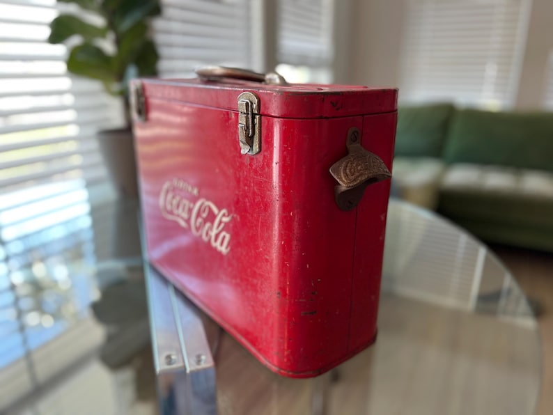 K&ouml;nnte beinhalten: Vintage rote Coca-Cola-K&uuml;hltasche mit Metallgriff und Flaschen&ouml;ffner. Das Coca-Cola-Logo ist in wei&szlig;er Schrift auf der Seite zu sehen. Die rechteckige K&uuml;hltasche scheint aus Metall zu sein.