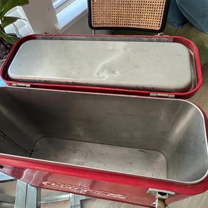 K&ouml;nnte beinhalten: Ge&ouml;ffnete rote Metallk&uuml;hlbox mit silbernem Innenraum und Deckel. Die K&uuml;hlbox hat eine rechteckige Form mit abgerundeten Ecken und Metallverschl&uuml;ssen. Der Deckel ist ge&ouml;ffnet und gibt den Innenraum frei. Die K&uuml;hlbox ist wahrscheinlich vintage.