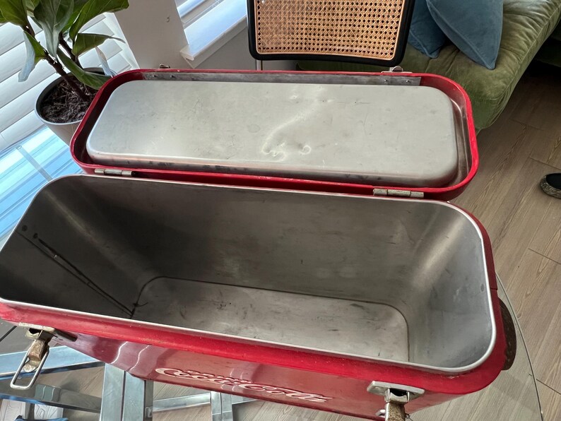 K&ouml;nnte beinhalten: Ge&ouml;ffnete rote Metallk&uuml;hlbox mit silbernem Innenraum und Deckel. Die K&uuml;hlbox hat eine rechteckige Form mit abgerundeten Ecken und Metallverschl&uuml;ssen. Der Deckel ist ge&ouml;ffnet und gibt den Innenraum frei. Die K&uuml;hlbox ist wahrscheinlich vintage.