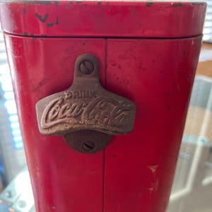 K&ouml;nnte beinhalten: Vintage rote Coca-Cola-K&uuml;hltruhe mit einem Metallflaschen&ouml;ffner. Der &Ouml;ffner ist vorne angebracht und tr&auml;gt das Coca-Cola-Logo und das Wort "DRINK" als Pr&auml;gung. Die K&uuml;hlbox weist Gebrauchsspuren auf.