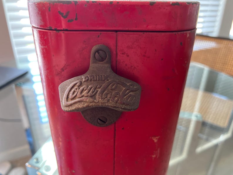 K&ouml;nnte beinhalten: Vintage rote Coca-Cola-K&uuml;hltruhe mit einem Metallflaschen&ouml;ffner. Der &Ouml;ffner ist vorne angebracht und tr&auml;gt das Coca-Cola-Logo und das Wort "DRINK" als Pr&auml;gung. Die K&uuml;hlbox weist Gebrauchsspuren auf.