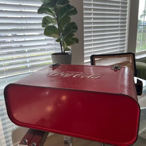 K&ouml;nnte beinhalten: Vintage rote Coca-Cola-K&uuml;hltasche mit wei&szlig;em Logo auf dem Deckel. Die K&uuml;hltasche hat Metallverschl&uuml;sse und eine rechteckige Form. Die K&uuml;hltasche steht auf einem Glastisch mit Chromgestell.
