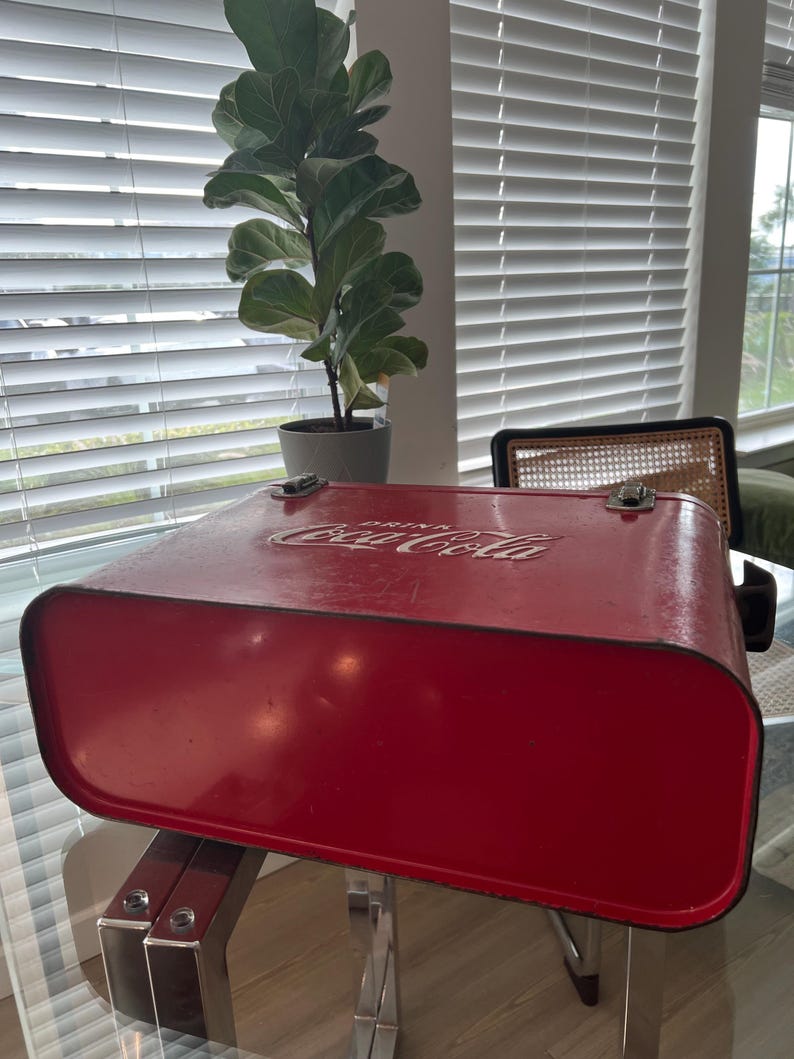 K&ouml;nnte beinhalten: Vintage rote Coca-Cola-K&uuml;hltasche mit wei&szlig;em Logo auf dem Deckel. Die K&uuml;hltasche hat Metallverschl&uuml;sse und eine rechteckige Form. Die K&uuml;hltasche steht auf einem Glastisch mit Chromgestell.