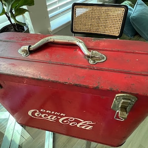 K&ouml;nnte beinhalten: Vintage rote Coca-Cola-K&uuml;hltasche mit silbernem Griff und Verschl&uuml;ssen. Das Coca-Cola-Logo in Wei&szlig; ist auf der Seite zu sehen. Die rechteckige K&uuml;hltasche weist Gebrauchsspuren auf.