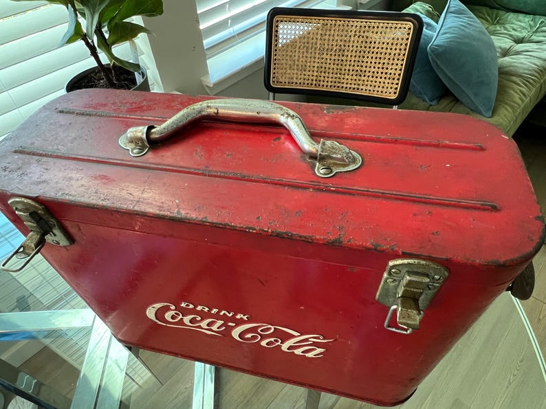 K&ouml;nnte beinhalten: Vintage rote Coca-Cola-K&uuml;hltasche mit silbernem Griff und Verschl&uuml;ssen. Das Coca-Cola-Logo in Wei&szlig; ist auf der Seite zu sehen. Die rechteckige K&uuml;hltasche weist Gebrauchsspuren auf.