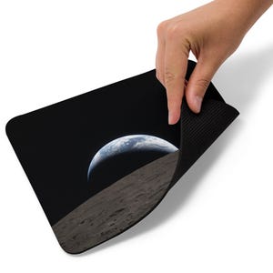 Puede incluir: Alfombrilla de ratón rectangular negra con una imagen de la Tierra elevándose sobre la superficie lunar sobre un fondo negro. La alfombrilla se levanta, revelando la parte inferior negra y texturizada. Tema espacial.