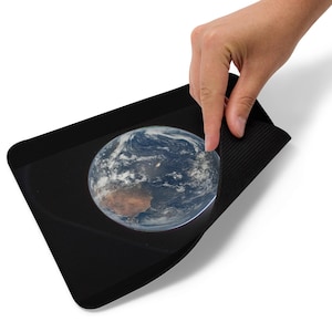 May include: A black rectangular mousepad featuring a detailed image of Earth. The image shows the continents, oceans, and clouds of the planet. A hand is lifting the edge of the mousepad.