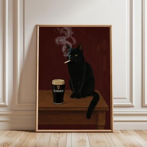 K&ouml;nnte beinhalten: Gerahmter Kunstdruck mit einer schwarzen Katze, die eine Zigarette raucht, mit einem Pint Guinness daneben. Die Katze sitzt auf einem Holztisch vor einem dunkelroten Hintergrund. Das Guinness-Glas zeigt das Markenlogo.
