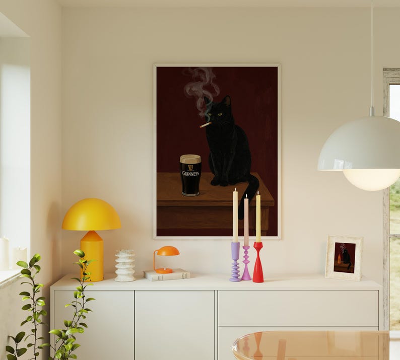 K&ouml;nnte beinhalten: Ein gerahmter Kunstdruck zeigt eine schwarze Katze, die eine Zigarette raucht, mit einem Pint Guinness auf einer Holzoberfl&auml;che. Der Hintergrund ist tiefrot. Der Druck wird &uuml;ber einem wei&szlig;en Schrank mit Kerzen und einem kleinen gerahmten Bild angezeigt.