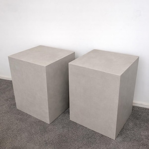 May include: Two light gray, cube-shaped pedestals. These modern, minimalist pedestals are made of a solid material and are suitable for displaying art, plants, or other decorative items. The pedestals are placed on a gray carpet.