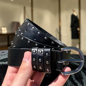 Puede incluir: Un cinturón de cuero negro con tachuelas plateadas y una hebilla plateada. La imagen muestra un primer plano del cinturón, destacando el diseño tachonado. Un accesorio elegante.