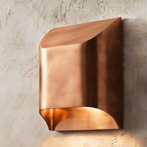 Op de afbeelding: Een wandlamp met een gebogen, metalen buitenkant in koperkleur. Het ontwerp heeft een glad, massief voorpaneel dat aan de bovenkant licht naar buiten helt, met een open onderkant die een warm, neerwaarts gericht licht verspreidt.