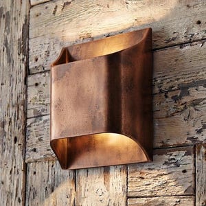Puede incluir: Un aplique de pared rectangular con acabado en cobre envejecido. La luminaria presenta un diseño curvo abierto en ambos extremos, que dirige la luz hacia arriba y hacia abajo, creando un brillo cálido sobre la superficie de montaje.