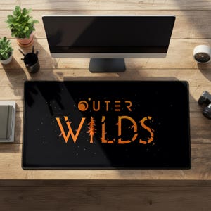 Pode incluir: Um tapete de secretária retangular preto com o texto laranja Outer Wilds no centro. O design inclui um ícone de planeta laranja estilizado acima da palavra Outer e uma silhueta de árvore integrada na letra I da palavra Wilds.