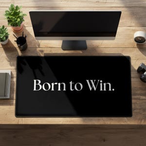 Peut inclure: Un tapis de bureau rectangulaire noir avec le texte blanc Born to Win. centré sur sa surface. Le tapis est placé sur un bureau en bois devant un écran d'ordinateur, avec une petite plante en pot, un porte-stylo et une horloge à proximité.