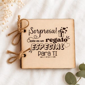 以下が含まれることがあります： 「¡Sorpresa! Este es un regalo ESPECIAL Para TI」と黒字で書かれた正方形の木製ギフトタグ。タグはひもで結ばれており、小さなギフトボックスとハートのイラストが付いています。背景は白い布です。