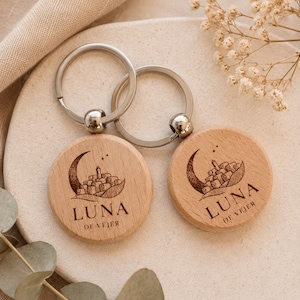 Puede incluir: Dos llaveros circulares de madera con un diseño grabado a láser de una luna creciente que acuna un pequeño pueblo. El texto LUNA DE VEJER está grabado debajo de la ilustración. Cada llavero incluye una anilla metálica plateada.