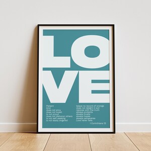 May include: A framed teal and white print featuring the word "LOVE" in large, bold letters. The print includes a list of qualities associated with love and the reference "1 Corinthians 13". The frame is black and the print is displayed on a wooden floor.