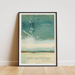 May include: Framed art print with a teal and beige abstract background. The word "love" is written in cursive. The text "Love Never Fails" and a Bible verse are at the bottom. The artwork is displayed on a wooden floor.