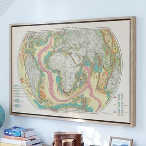 May include: A framed geological world map hanging on a white wall above two wooden dressers. The map features colorful tectonic plate boundaries and landmasses. A globe, a stack of books, a brown leather bag, and a small framed photo sit on top.