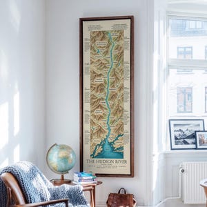 May include: A tall, vertical framed map of the Hudson River, featuring detailed topographical illustrations of the surrounding mountains and towns, with the title The Hudson River printed in bold black text at the bottom of the cream-colored map.