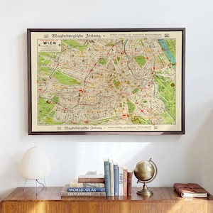 May include: A framed vintage map of Vienna titled Pharos-Plan Wien, featuring detailed street layouts, green park areas, and red transit lines on a cream background, enclosed in a dark brown wooden frame and displayed on a wall.